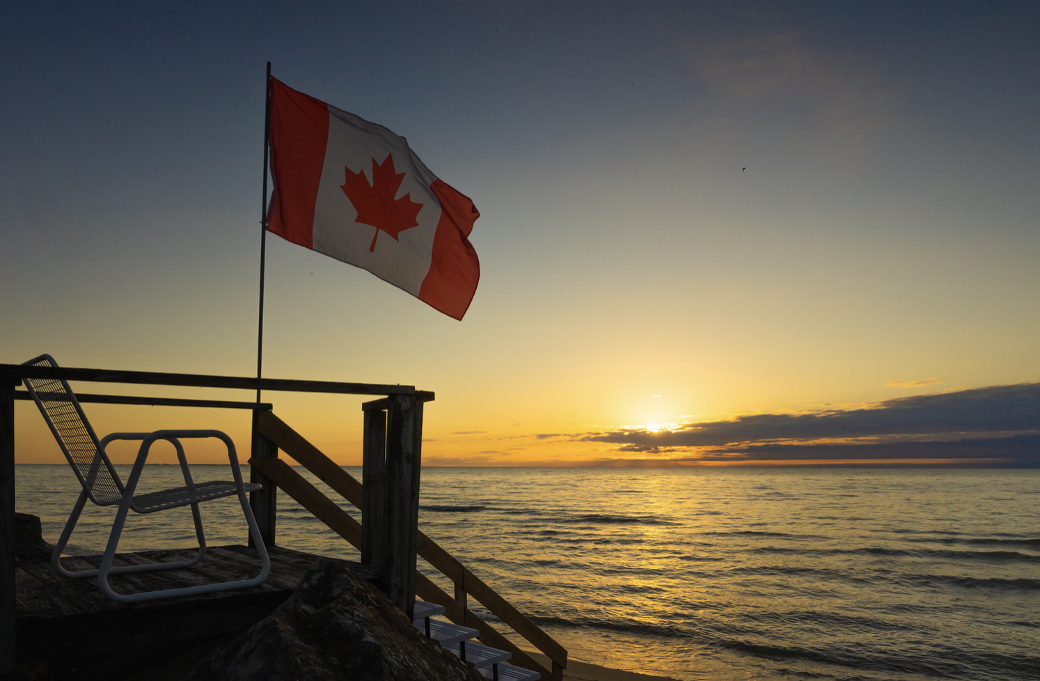 Photo of Canadian flag blowing in breeze over Lake Erie next to an outdoor lounge chair. Photo of Canadian flag blowing in breeze over Lake Erie next to an outdoor lounge chair.